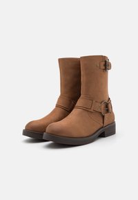 Botines de ante marrón hasta el tobillo con puntera redonda, que presentan un detalle decorativo de hebilla y correa, y un tacón bajo y robusto.