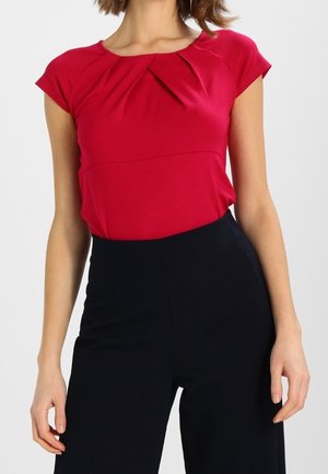 Vrouw draagt een rood kort mouwblouse met plooien bij de hals, gestopt in een zwarte broek met hoge taille en wijde pijpen.