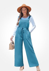 Teal overall jumpsuit met een gestrikt middel, wijde pijpen en voorzakken, gecombineerd met een witte lange mouw top en een bruine hoed met brede rand.