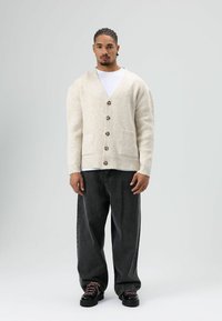 Beige wollen vest met een V-hals, knoopsluiting aan de voorkant en twee steekzakken. Gecombineerd met brede zwarte spijkerbroek en zwarte schoenen met roze veters.