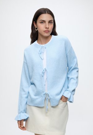 Femme aux longs cheveux foncés portant un cardigan tricoté bleu clair avec des liens de fermeture sur un haut blanc et une jupe crème sur fond uni.