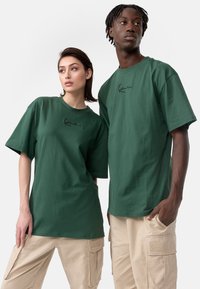 T-shirts en coton vert à manches courtes, ornés d'un petit logo noir sur la poitrine. Portés avec un pantalon cargo beige.
