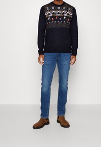 Granatowy sweter z geometrycznymi i świątecznymi wzorami, z okrągłym dekoltem i ściągaczami na rękawach. Połączony z niebieskimi dżinsami i brązowymi botkami.