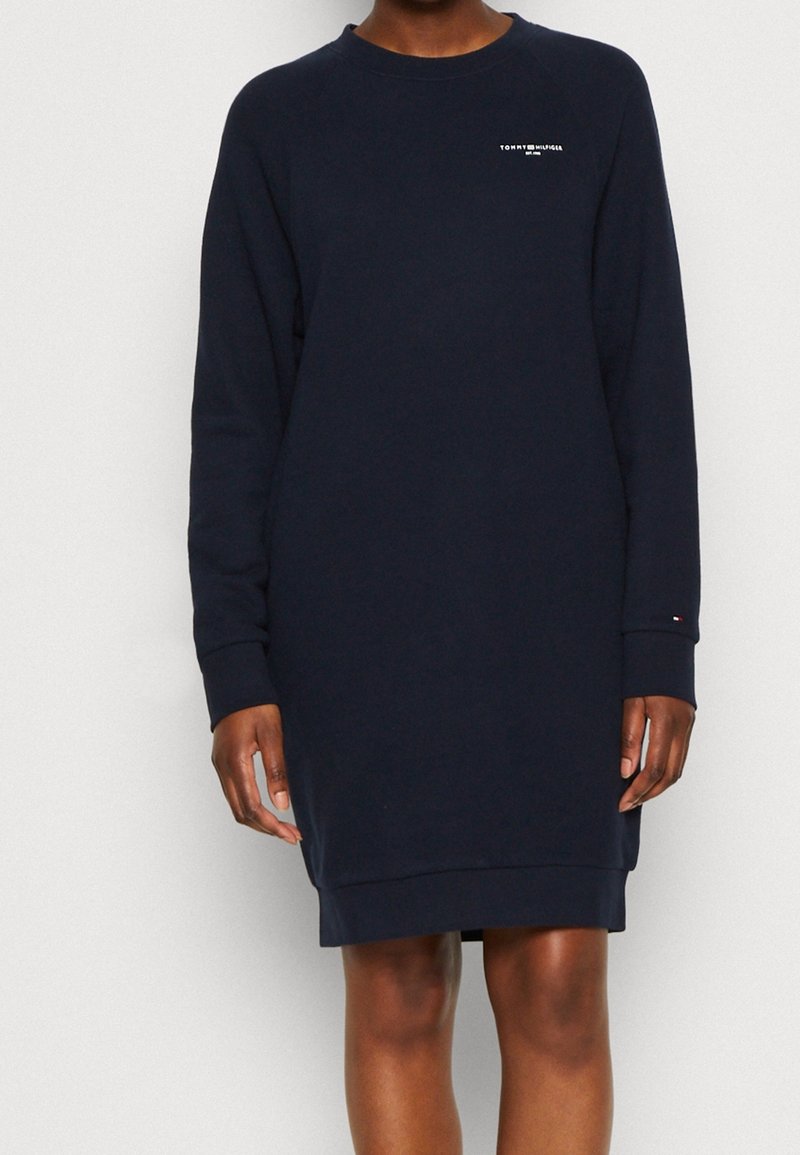 Personne portant une robe bleu marine à manches longues avec un petit logo blanc sur la poitrine et la manche, se tenant devant un fond uni clair.