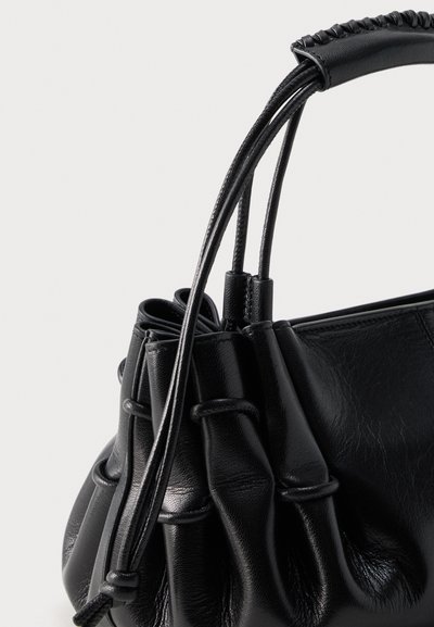 Μαύρη δερμάτινη τσάντα με μαζεμένες πλευρές, πλεγμένη λεπτομέρεια στη λαβή και λεπτές δερμάτινες λωρίδες σε απλό ανοιχτό φόντο.