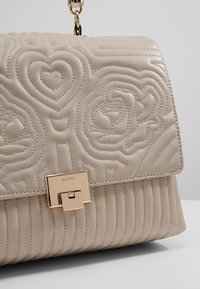 Sac à main en cuir beige avec motifs de cœur et de rose cousus, fermoir rectangulaire doré marqué "ALDO," et une bandoulière en chaîne dorée.