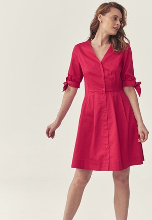 Kobieta ubrana w czerwoną sukienkę na guziki o długości do kolan z rękawami do połowy przedramienia związanymi kokardami, stojąca na jasnym tle.