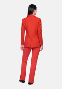 Roter maßgeschneiderter Blazer und passende Hose, strukturierter Stoff, schlanke Passform, mit einem einzelnen Knopfdetail auf der Rückseite und ausgestellten Hosenbeinen.