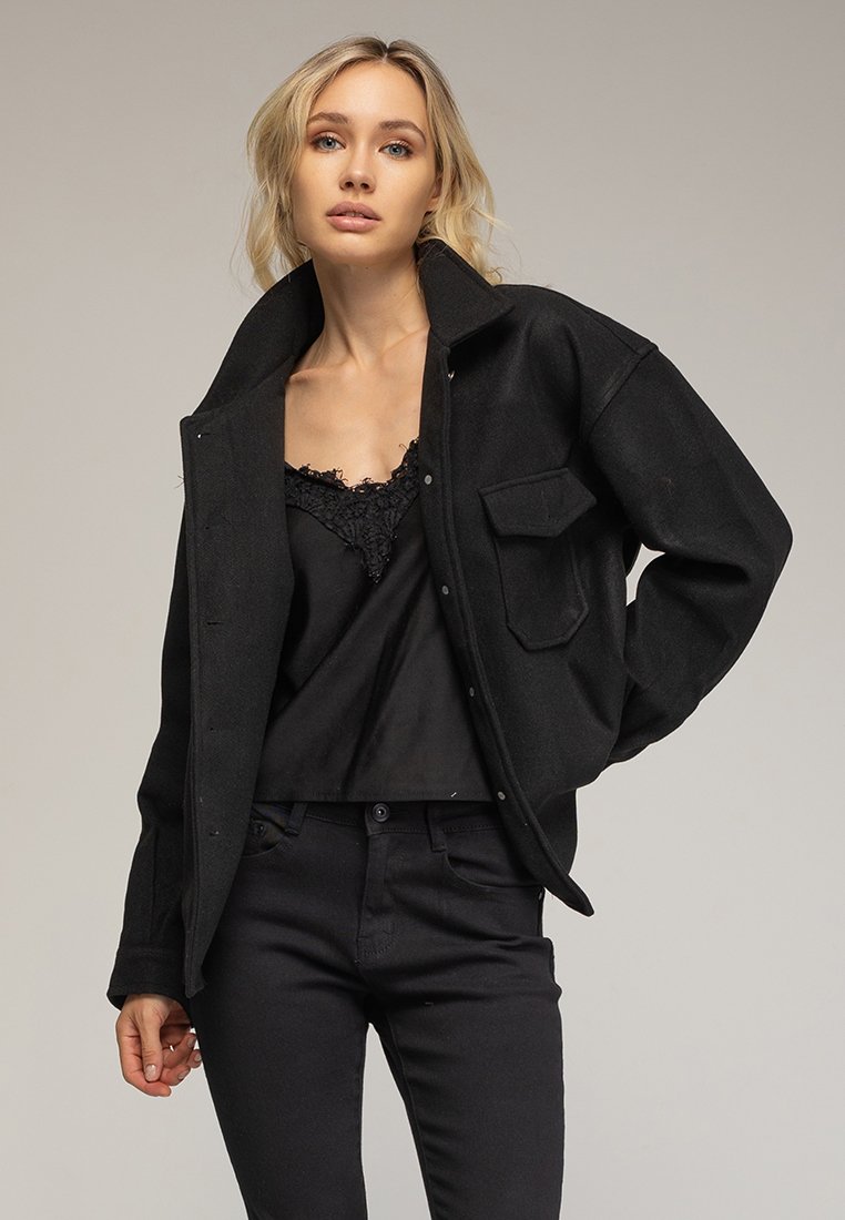 Chaqueta de mezcla de lana negra con un cuello, bolsillos frontales y cierre de botones, llevada sobre una blusa negra con encaje y pantalones ajustados negros.