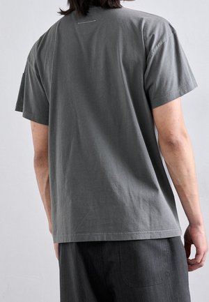 Osoba widziana od tyłu, ubrana w prostą ciemnoszarą koszulkę z krótkim rękawem i ciemne spodnie na jasnoszarym tle.