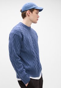 Blauwe kabelgebreide trui met een hoge hals en ribbels aan de mouwen, gedragen over een wit shirt. Gecombineerd met een lichtblauwe pet, zijaanzicht weergegeven.