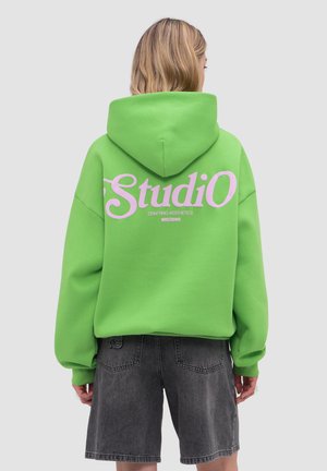 Person trägt leuchtend grünen Hoodie mit großem blassrosa Schriftzug "Studio" auf dem Rücken, kombiniert mit dunkelgrauen Denim-Shorts.