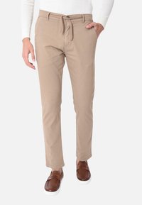 Pantaloni beige con vita regolabile, due tasche laterali e vestibilità affusolata. Indossati con mocassini marroni, su uno sfondo neutro.
