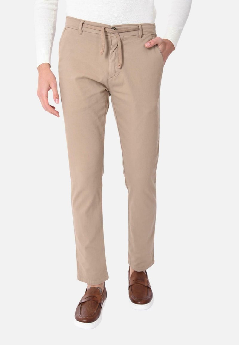Pantaloni beige con vita regolabile, due tasche laterali e vestibilità affusolata. Indossati con mocassini marroni, su uno sfondo neutro.