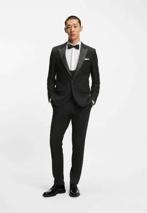 Tuxedo nero con rever in satin, camicia bianca e papillon nero. Pantaloni aderenti, scarpe nere lucide, design minimalista.