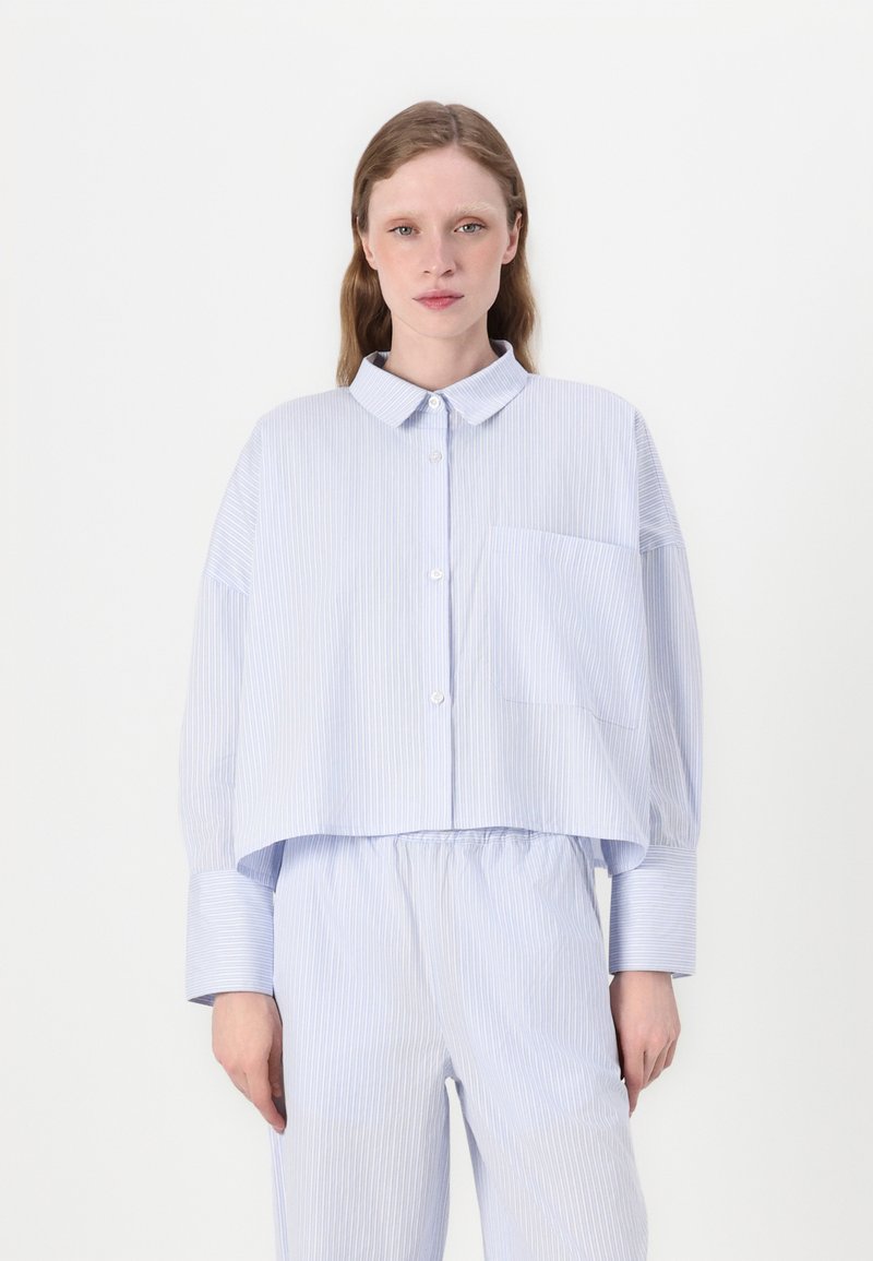 Camisa curta azul e branca com riscas, com colarinho, mangas longas, um único bolso no peito e um corte descontraído, usada com calças a condizer.