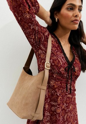 Frau trägt ein rotes Blumenkleid mit schwarzem Besatz und eine große beige Wildleder-Umhängetasche mit verstellbarem Riemen und goldener Schnalle.