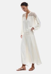 Robe blanche légère et longue avec un corsage texturé, des détails en dentelle sur les épaules, et une jupe fluide. Associée à des sandales brunes à lanières.