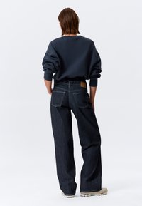 Sweat-shirt oversize bleu foncé avec poignets côtelés et jean en denim foncé ample assorti. Style décontracté avec une patch en cuir beige sur le jean.