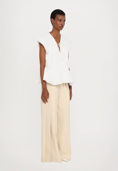 White peplum top with front tie detail and cap sleeves, paired with beige wide-leg trousers. Soft fabric, relaxed fit, minimalistic design.