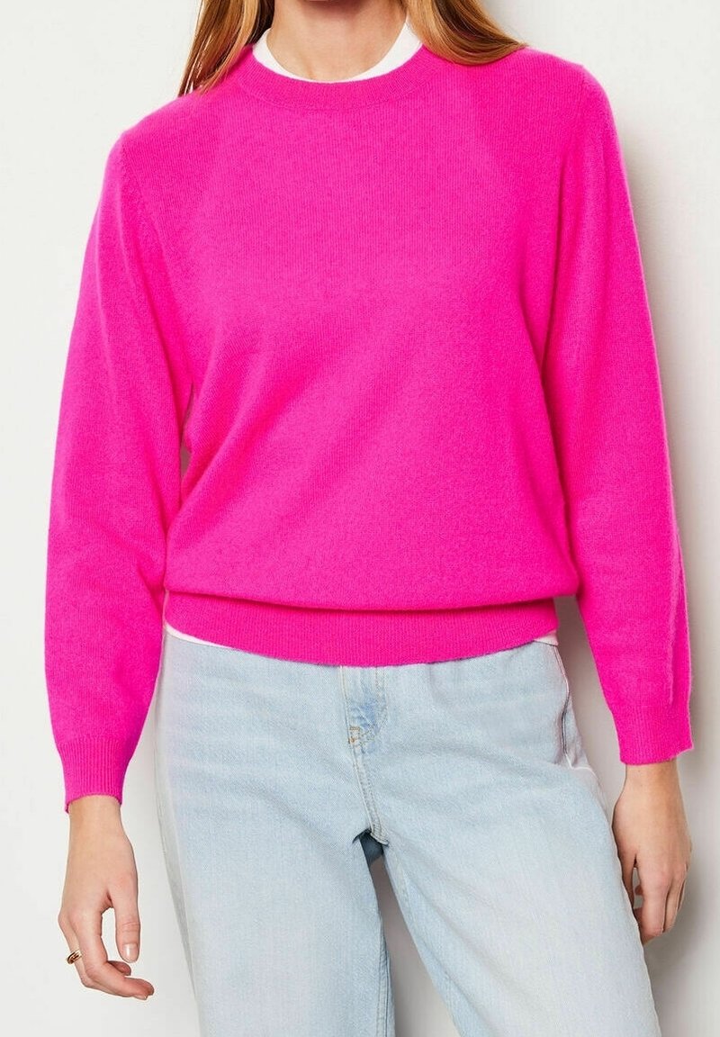 Femme portant un pull manches longues rose vif sur un sous-pull blanc, associé à un jean bleu clair, debout devant un fond neutre.