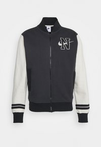 Nike-takki, jossa on musta vartalo, kerma väriset hihat, ribbausviimeistely ja kirjailtu logo. Vetoketjukiinnitys ja korostetut sivutaskut.