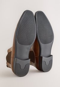 Braune Lederschuhe mit strukturierten grauen Gummisohlen. Die Sohlen haben ein gemustertes Profil für besseren Halt und ein offenes Fersendesign.