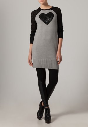 Graues Strickkleid mit schwarzen Netzärmeln, verziert mit einem schwarzen Paillettenherz-Design. Gestylt mit schwarzen Leggings und schwarzen High Heels.