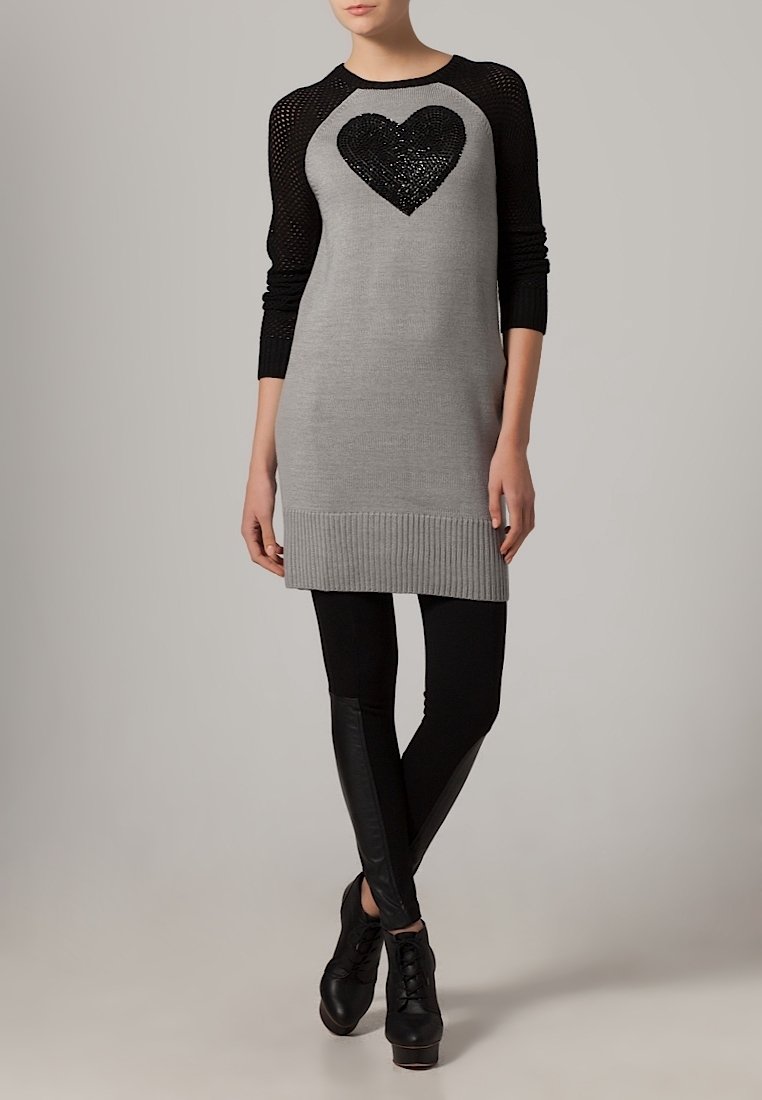 Robe en tricot grise avec des manches en maille noires, ornée d'un design en sequins noirs en forme de cœur. Associée à des leggings noirs et des chaussures à talons noires.