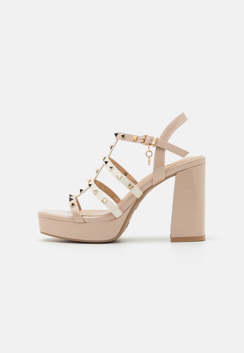 Sandalias de plataforma beige con correas blancas, herrajes dorados y acentos de tachuelas negras. Tacón alto grueso y textura suave.