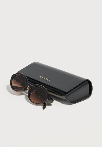 Στρογγυλά γυαλιά ηλίου με καφέ ταρταρούγα και σκουρούς φακούς δίπλα σε μαλακή μαύρη θήκη με ανάγλυφο το λογότυπο "GUCCI" σε χρυσά γράμματα.