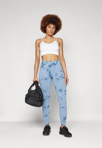 Crop top de sport blanc avec détail froncé, leggings bleu clair à motif marbré, sac de gym noir et chaussures de sport noires.