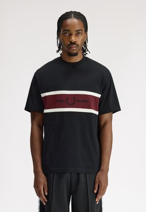 Homme aux cheveux tressés portant un T-shirt noir avec des rayures bordeaux et blanches et le logo "Fred Perry", debout sur fond blanc.