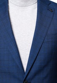 Granatowa marynarka w kratę z cienkimi niebieskimi liniami, na dwa guziki, z czarnym akcentem na klapie, nałożona na jasnoszary sweter z golfem.