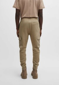 Beige Cargo-Hosen mit einem elastischen Bund, seitlichen Taschen und schmal zulaufenden Bündchen, kombiniert mit einem hellbraunen Textilhemd und braunen Stiefeln.