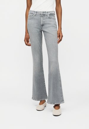 Személy magasderekú világosszürke bővülő farmert és bézs slip-on cipőt viselve, fehér háttér előtt áll.