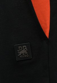 Μαύρο ύφασμα με πορτοκαλί εσωτερική επένδυση και μαύρο τετράγωνο έμβλημα που απεικονίζει έναν ταύρο σε επίθεση, τα γράμματα "PB" και τη λέξη "HUGO".