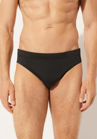 Schwarze Herren-Schwimmbriefe aus glattem Stoff, mit einem breiten Bündchen und minimalen Nähten. Kleines Logo-Detail an der Seite.