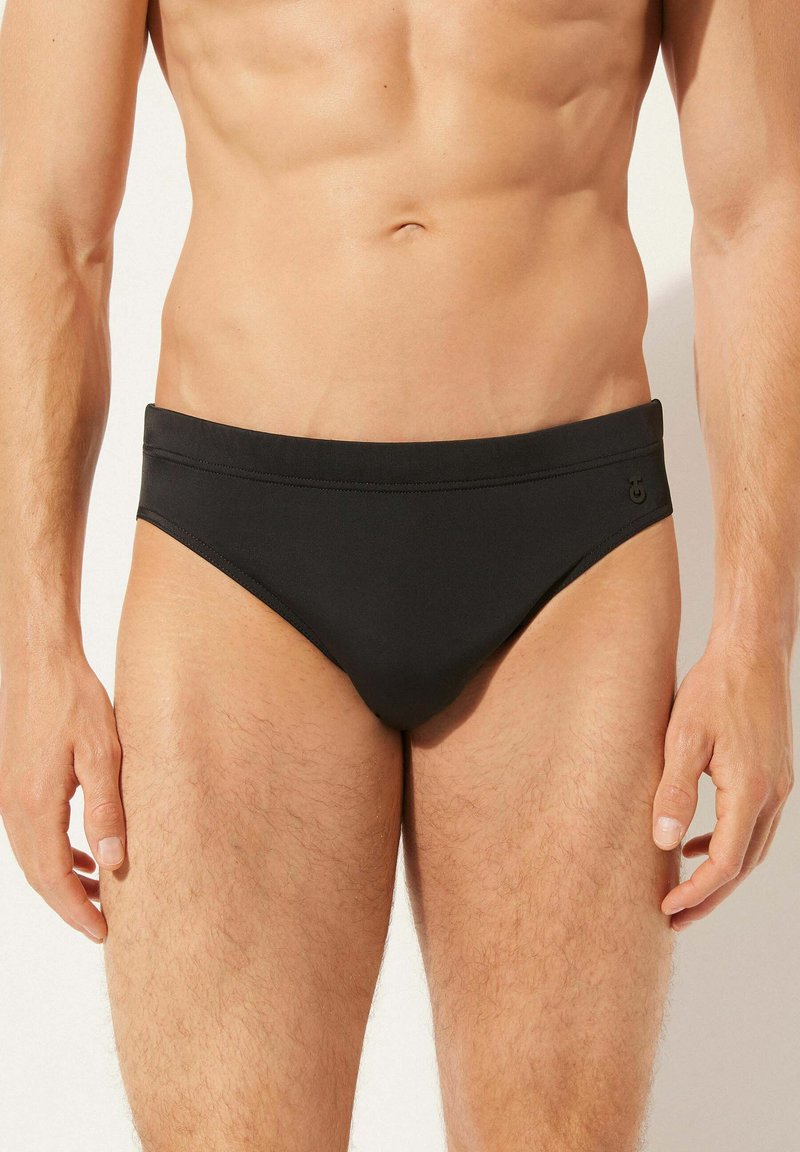 Schwarze Herren-Schwimmbriefe aus glattem Stoff, mit einem breiten Bündchen und minimalen Nähten. Kleines Logo-Detail an der Seite.