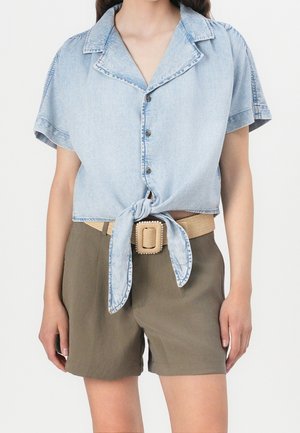 Vrouw draagt een lichtblauw denim shirt met korte mouwen, gestrikt in de taille met knopen, kaki korte broek en een beige riem met een vierkante gesp.