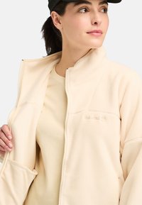 Light beige zip-up fleece jacket with a high collar, textured fabric, and subtle logo detail on the chest. Worn over a matching top.