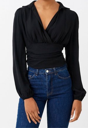 Frau trägt eine schwarze Wickelbluse mit langen Ärmeln und geraffter Taille, kombiniert mit hoch taillierten blauen Jeans.