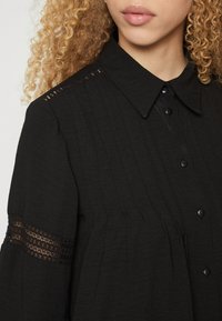 Μαύρο πουκάμισο με κουμπιά, χαρακτηριστικό του είναι ο γιακάς, η υφή του υφάσματος, οι ραφές pin-tuck και οι δαντέλες στα μανίκια.