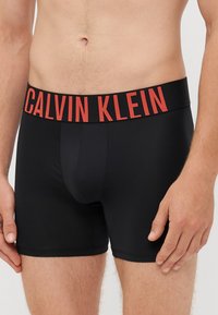 Boxeri negri din material elastic, cu un logo roșu "CALVIN KLEIN" pe talie, având o croială ajustată și picioare scurte.