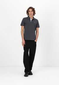 Mørk grå polo-skjorte med en kontrasterende lys grå krage, kombinert med svarte bukser og svarte sko. Modellen står foran en hvit bakgrunn.