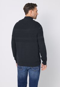 Pletený tmavě šedý svetr s vysokým límcem, zdobený texturovanými vodorovnými pruhy. Nošený s modrými džíny. Viditelné švy a žebrované manžety.