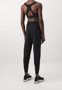 Schwarzer Sport-BH mit einem einzigartigen Rückenschlitz und strukturierten Trägern, kombiniert mit enganliegenden schwarzen Jogginghosen und schwarzen Sportschuhen.