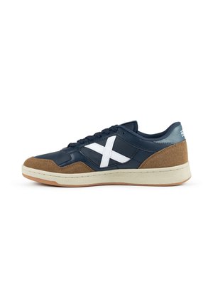 Zapatilla baja con tejido azul marino y detalles en ante marrón, logo blanco lateral en forma de cruz, suela crema y cordones negros sobre fondo blanco.