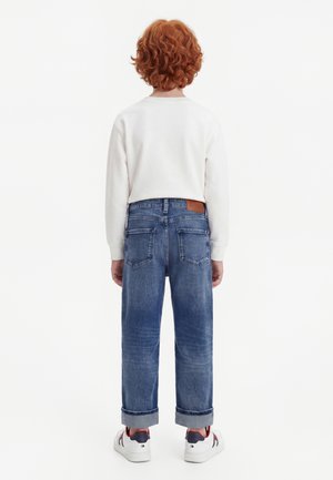 Barn med krøllet rødt hår iført en hvid langærmet trøje, blå opkrogede jeans og hvide sneakers, der står med ryggen til på en hvid baggrund.