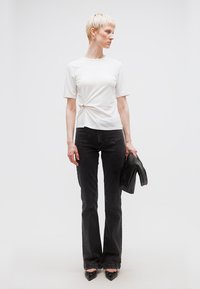 Γυναίκα με κοντά ξανθά μαλλιά που φοράει λευκό μπλουζάκι με πιέτες, μαύρο καμπάνα τζιν, μαύρα μυτερά παπούτσια, κρατώντας ένα μαύρο υφασμάτινο τσαντάκι.